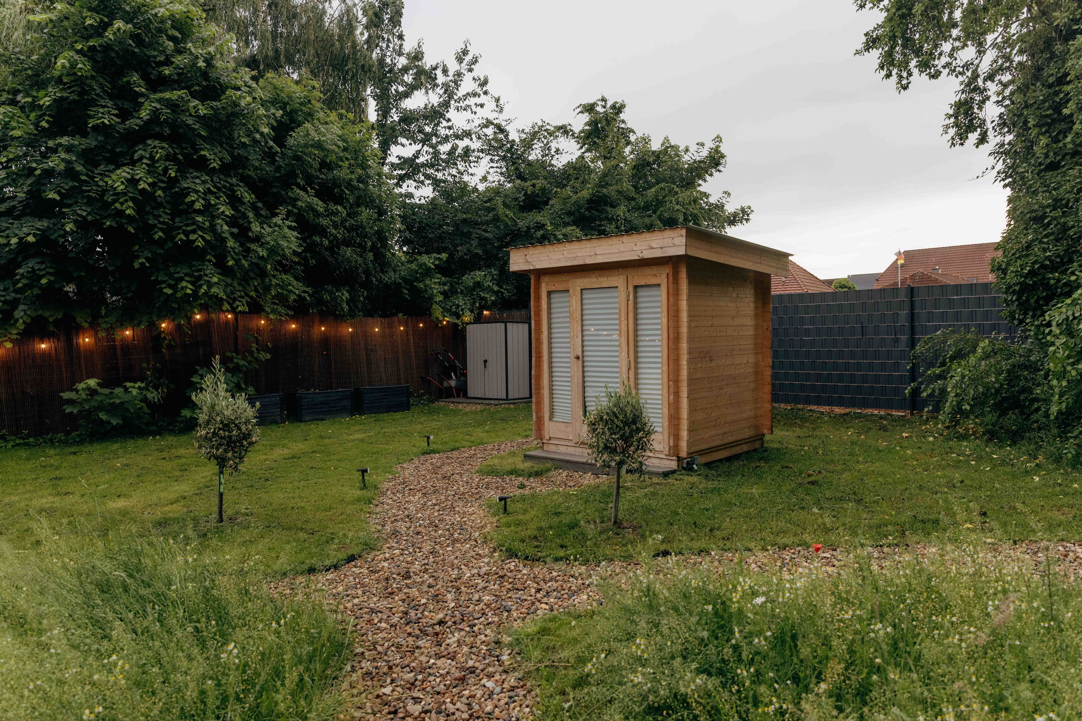 Freistehendes Badehaus aus Holz im Garten mit Kiesweg, Lichterkette und jungen Olivenbäumen – privates Bad fürs Bubble Zelt.